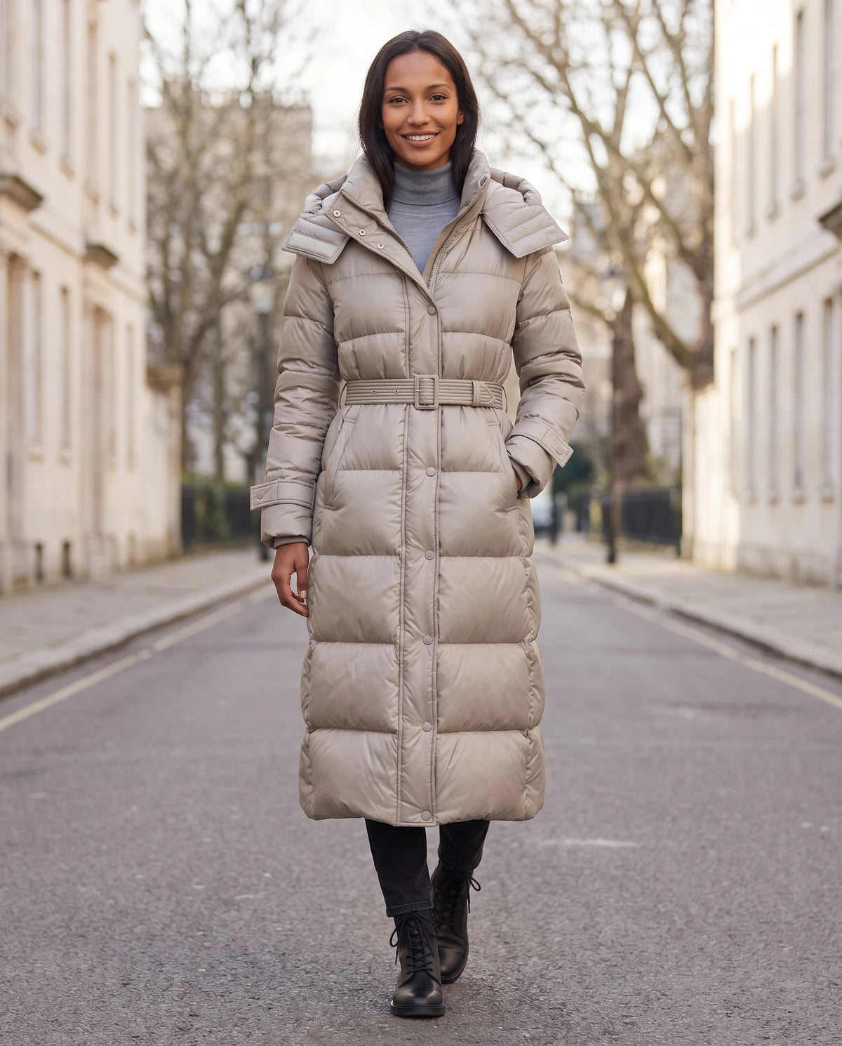 Extralange glänzende Steppjacke mit Kapuze und Schnallengürtel Carole in Beige, getragen auf einer Allee mit hellen Steingebäuden