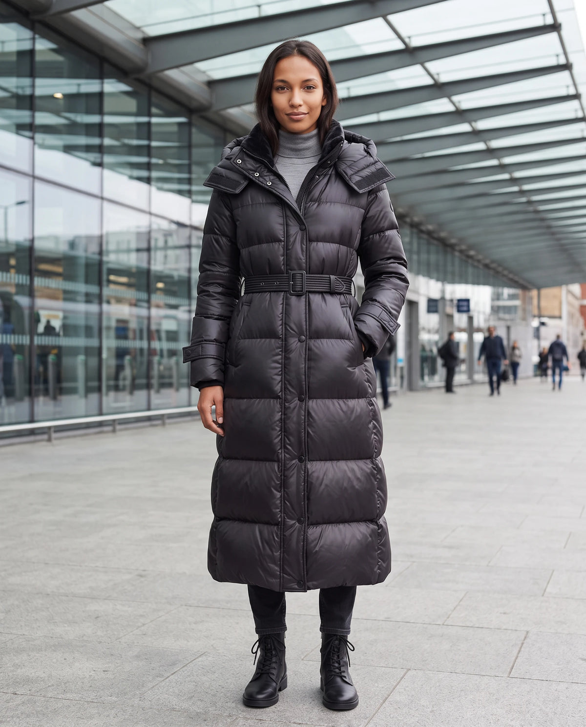 Extralange glänzende Steppjacke mit Kapuze und Schnallengürtel Carole in Schwarz, getragen unter dem Glasdach eines modernen Bahnhofsvorplatzes