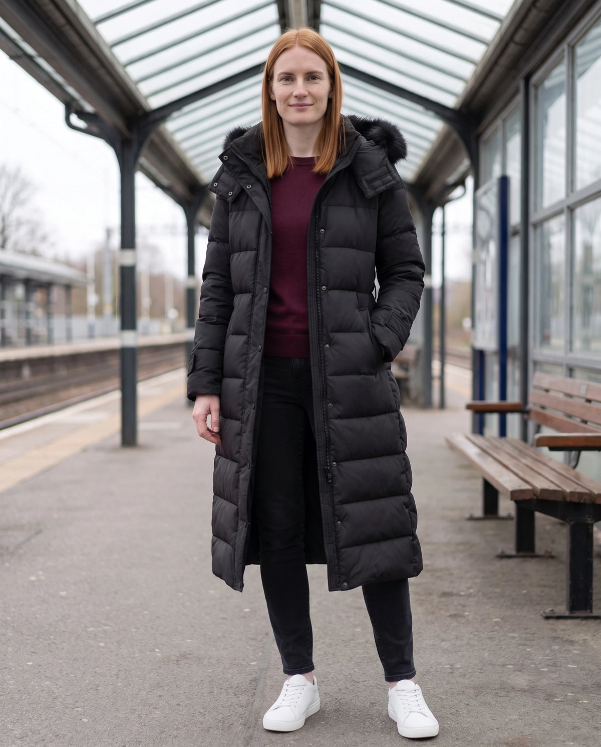 Doudoune extra-longue à capuche bordée et ceinture à nouer Élisabeth en Noir, portée sur un quai de gare couvert à la lumière douce