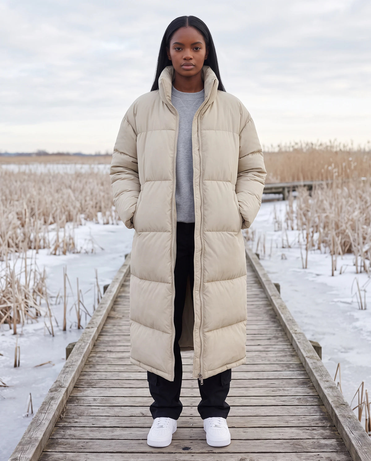 Extralanger Steppmantel mit Stehkragen und Zwei-Wege-Zip Cordélia in Beige, getragen auf einem Holzsteg über einem gefrorenen Moor