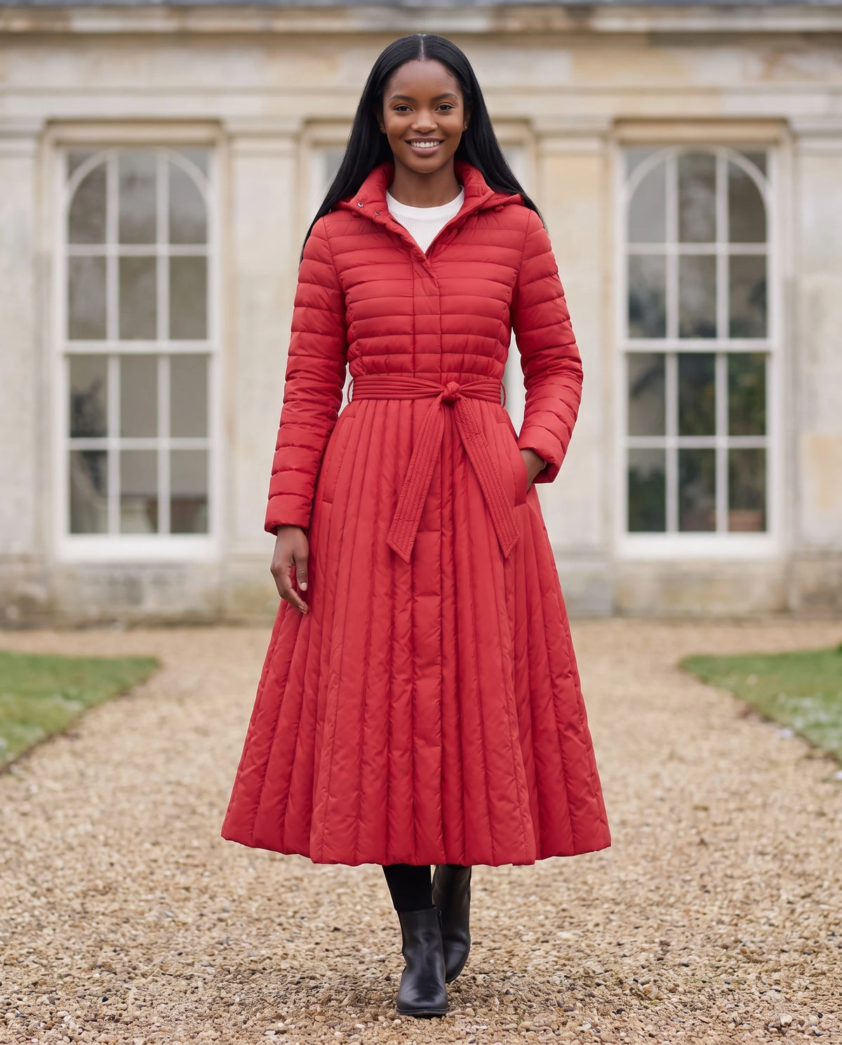 Doudoune extra-longue évasée à capuche et ceinture Tatiana en Rouge, portée devant une orangerie de pierre claire
