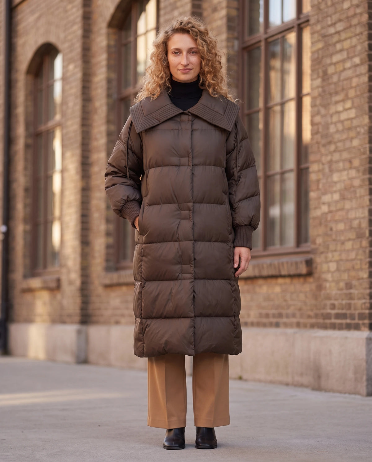 Doudoune extra-longue à grand col carré et poignets tricot Anastasia en Marron, portée devant une façade de brique brune