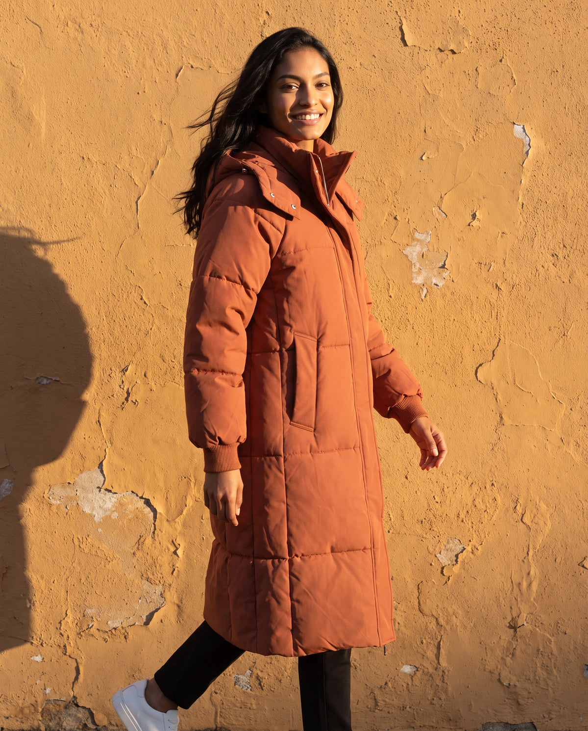 Doudoune longue à capuche Claire en Rouille, portée devant un mur ocre au soleil