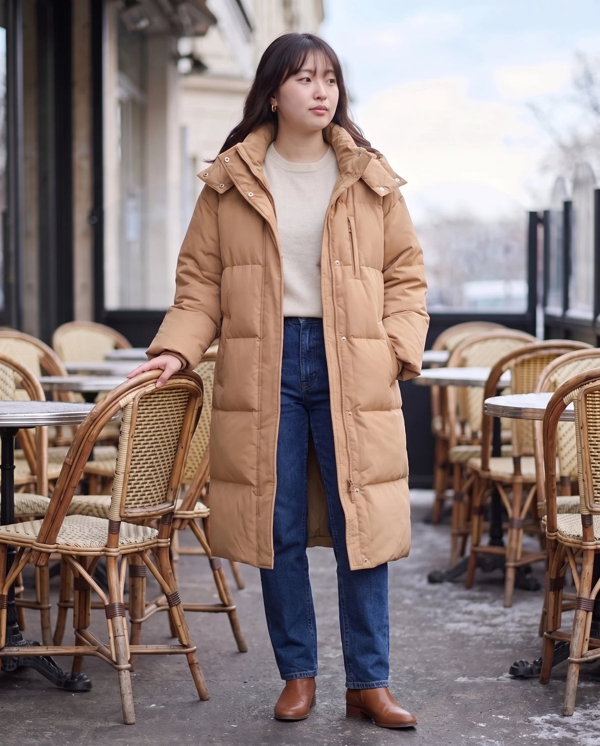 Langer Oversize-Steppmantel mit Kapuze Enora in Camel, getragen auf einer Winter-Caféterrasse