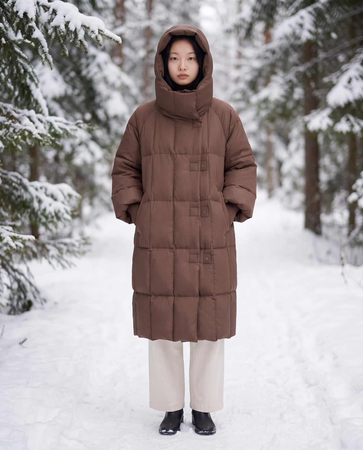 Doudoune longue oversize à col-capuche zippé Aloïse en Chocolat, portée capuche relevée sur un chemin forestier bordé de sapins enneigés