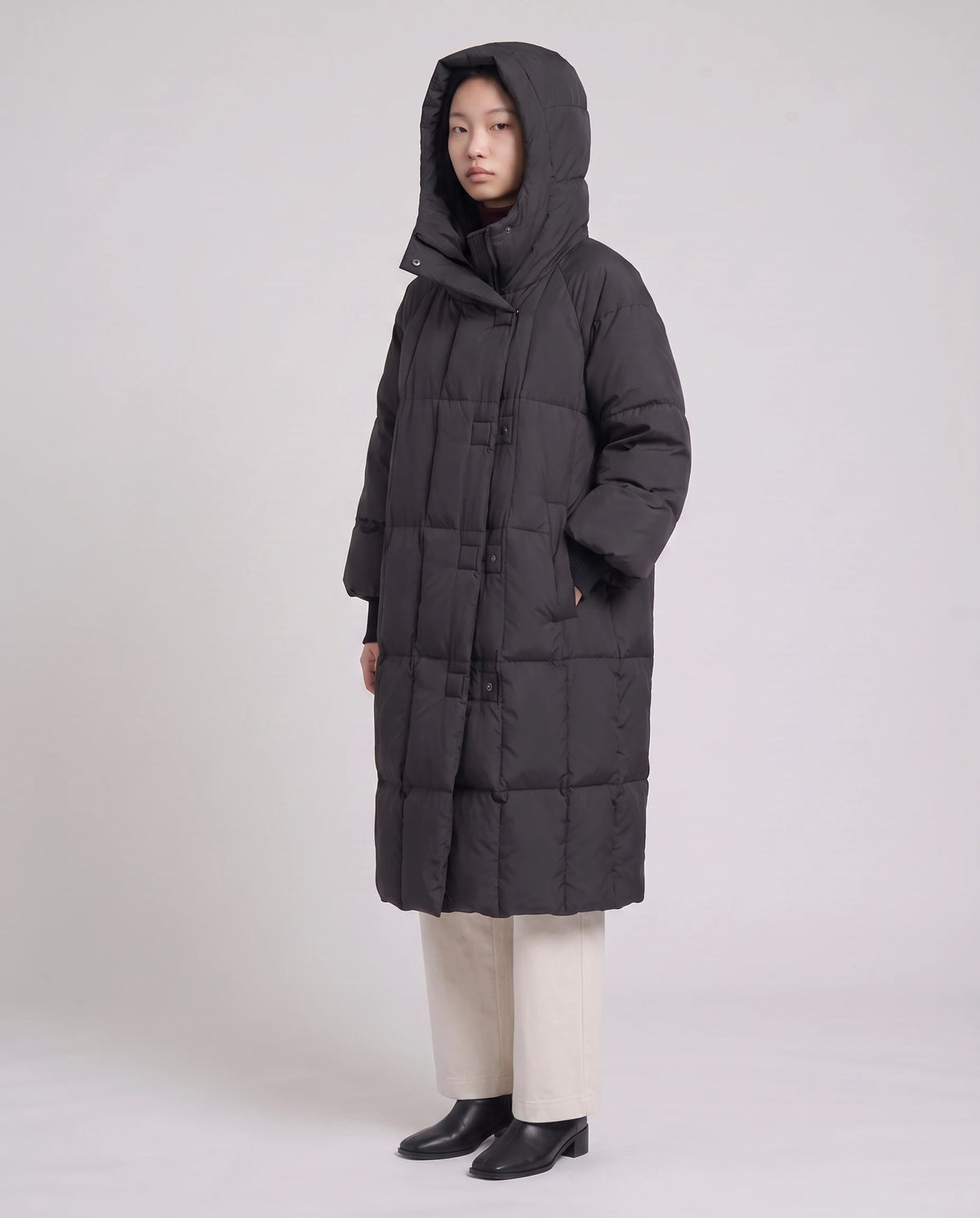 Doudoune longue oversize à col-capuche zippé Aloïse en Noir, vue de trois quarts, capuche zippée relevée, main sur le col