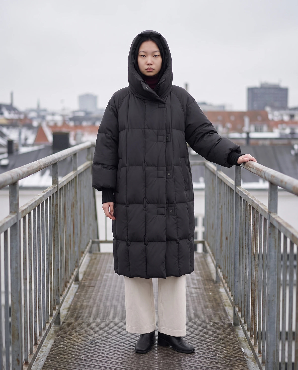 Doudoune longue oversize à col-capuche zippé Aloïse en Noir, portée capuche relevée sur une passerelle métallique sous un ciel gris