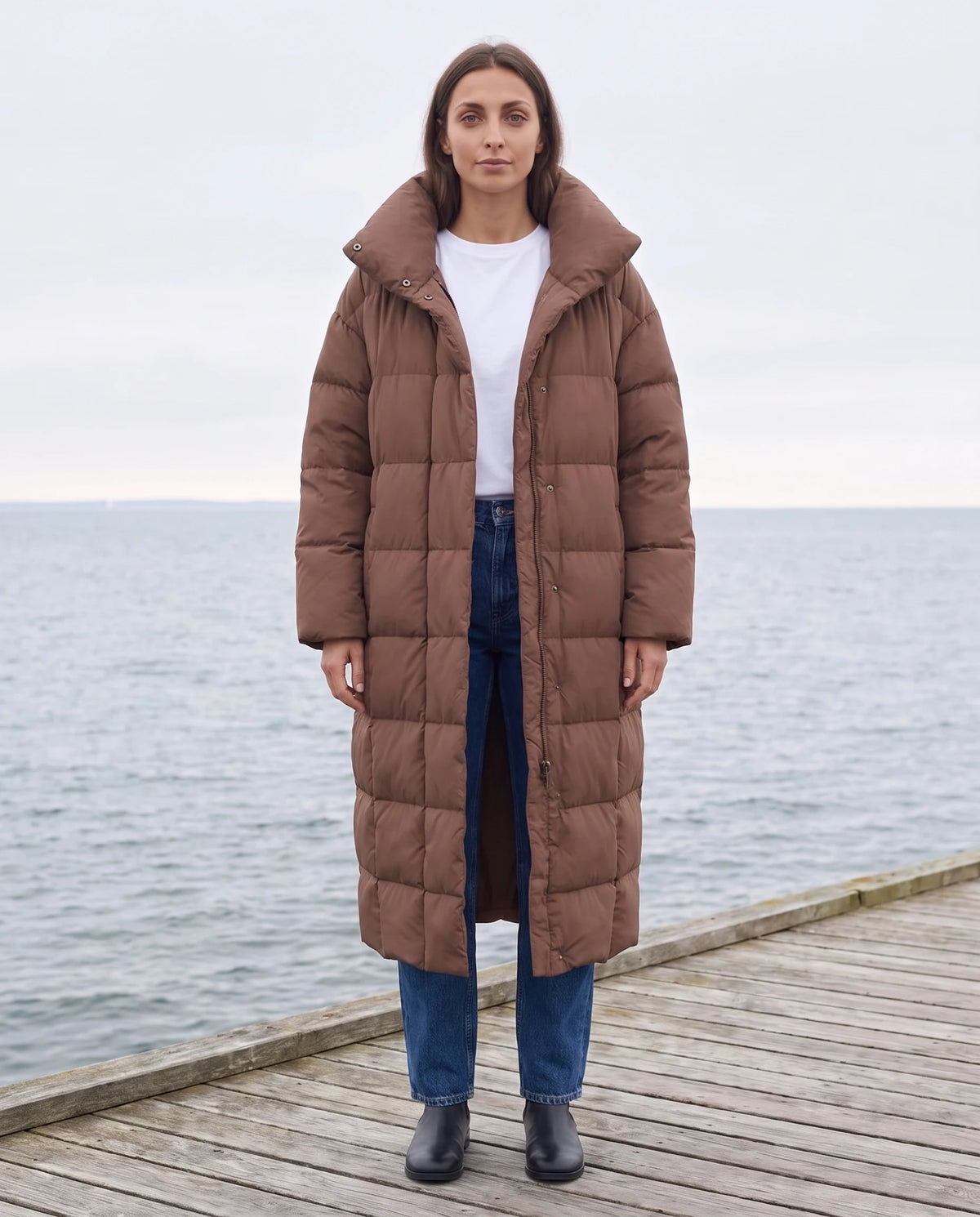 Doudoune longue oversize à col cheminée Laure en Chocolat, portée sur une jetée en bois face à la mer d'hiver, col entrouvert