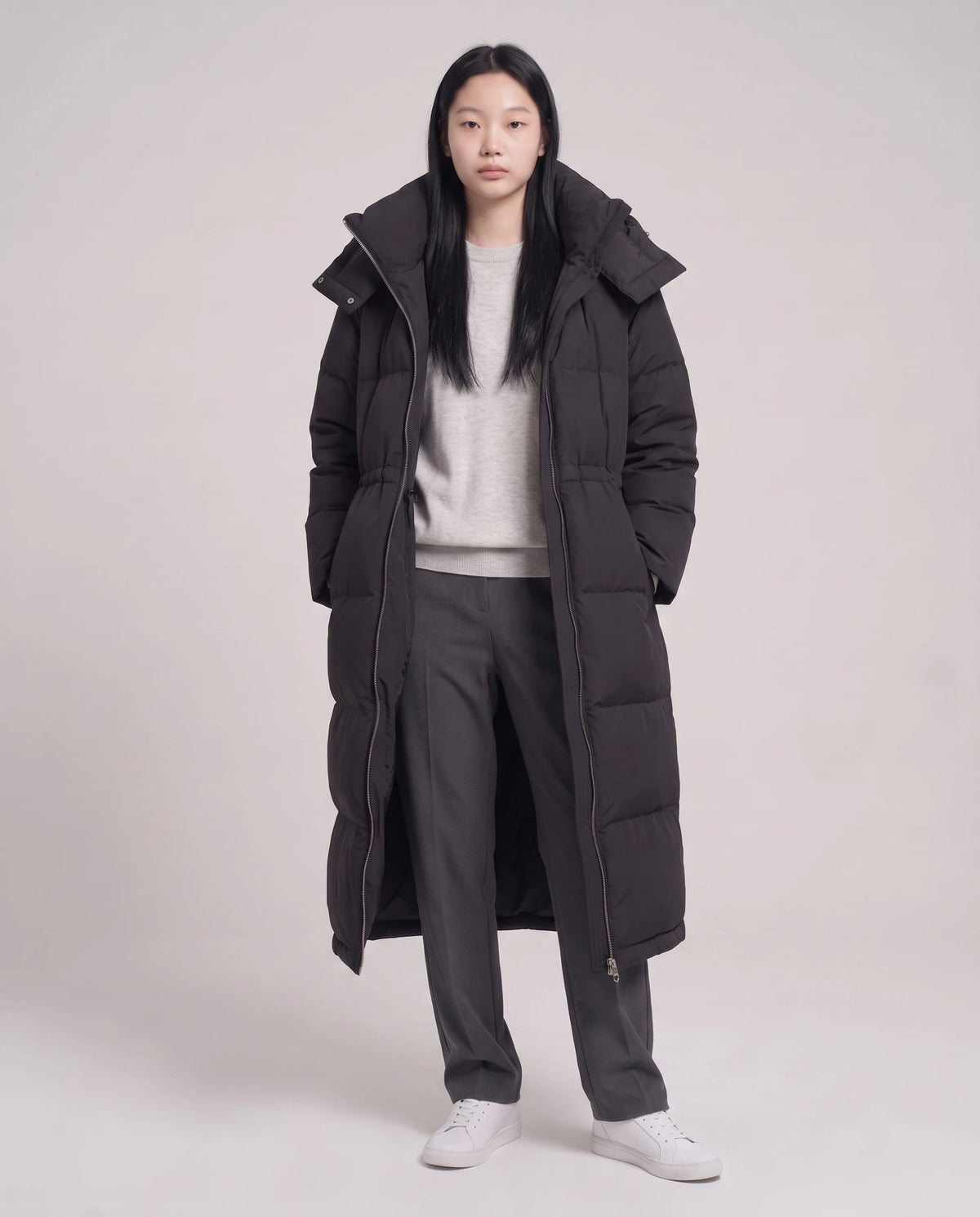 Langer Oversize-Steppmantel mit Stehkragen und Kapuze Lucille in Schwarz, offen getragen über graumeliertem Pullover, Kordel gelockert