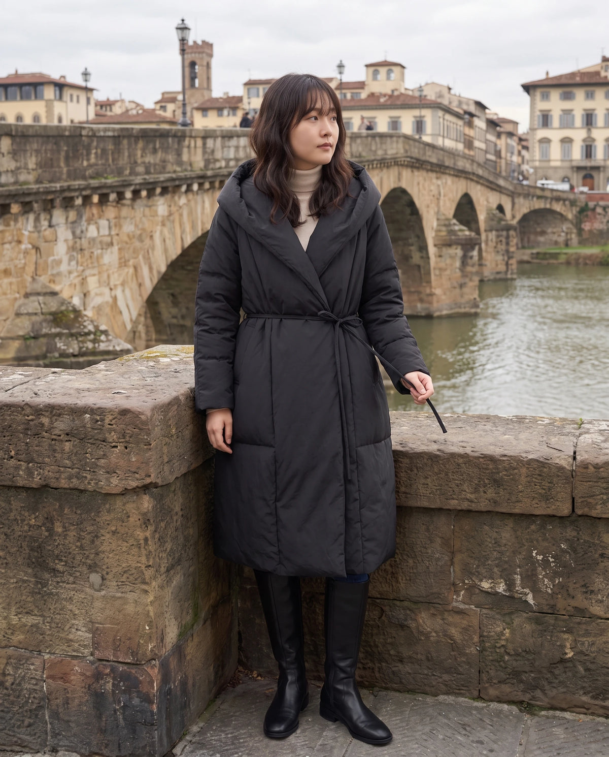 Doudoune longue portefeuille à capuche oversize Aurélia en Noir, portée sur un vieux pont de pierre au bord d'un fleuve
