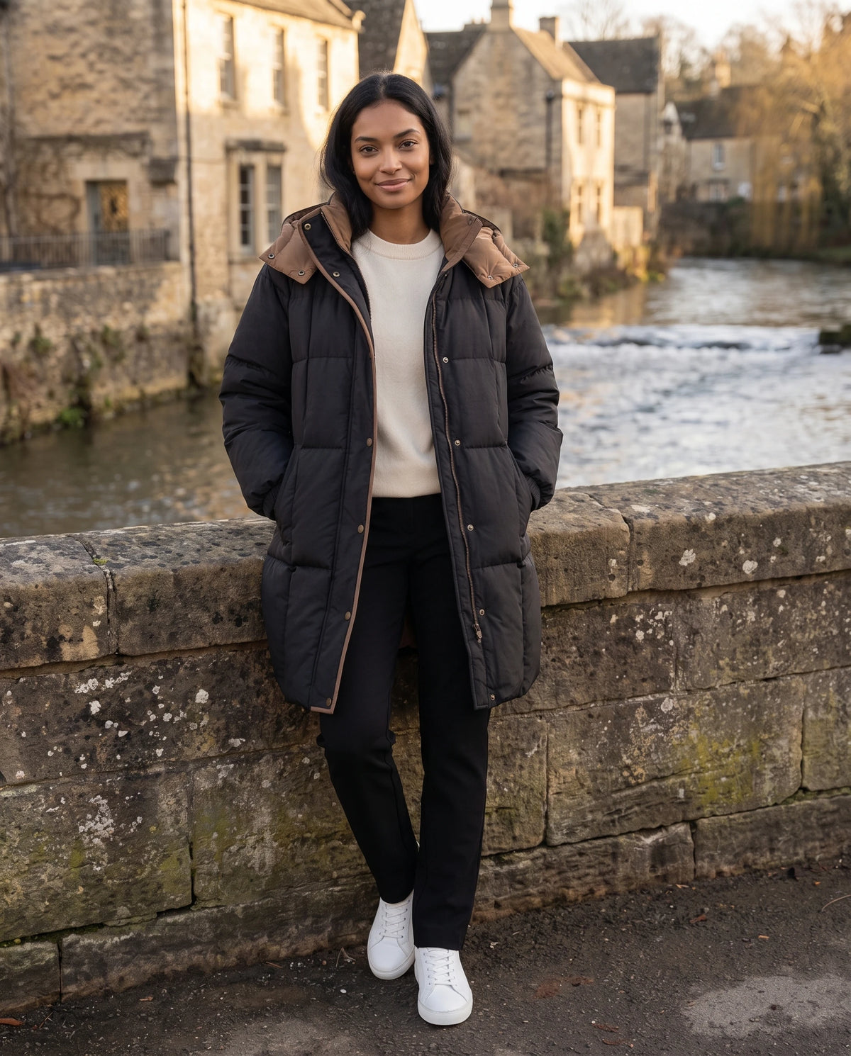 Mittellange Winterjacke mit Kapuze und Kontrastfutter Alexia in Schwarz, getragen an der Steinbrüstung einer Brücke