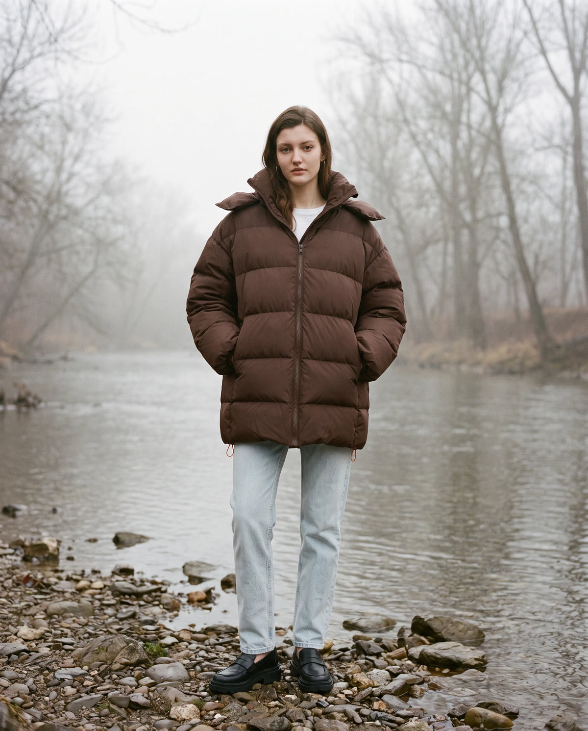 Oversize-Pufferjacke mit Kapuze Freya in Schokobraun, getragen an einem nebligen Flussufer