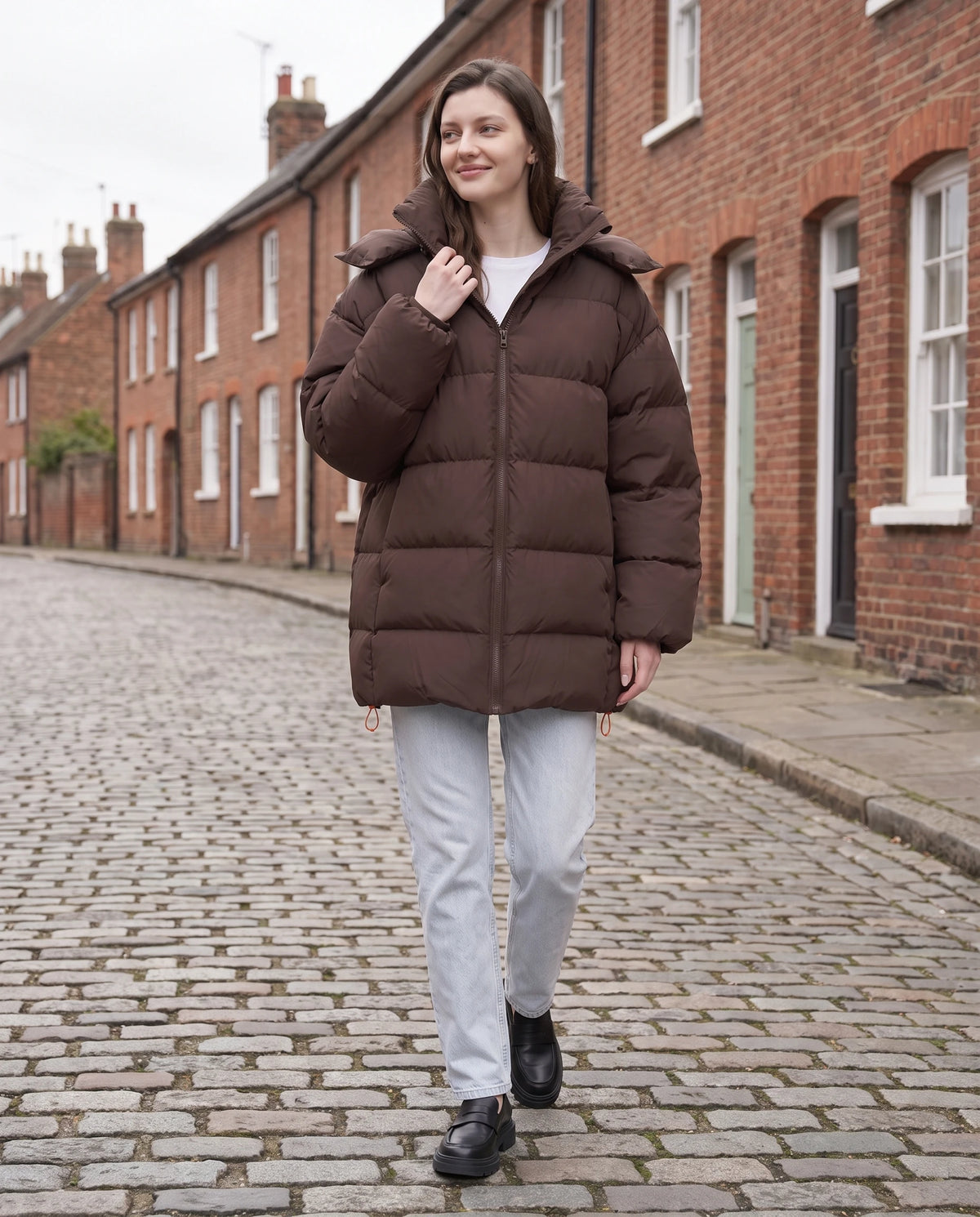Oversize-Pufferjacke mit Kapuze Freya in Schokobraun, getragen in einer Strasse mit roten Backsteinhäusern