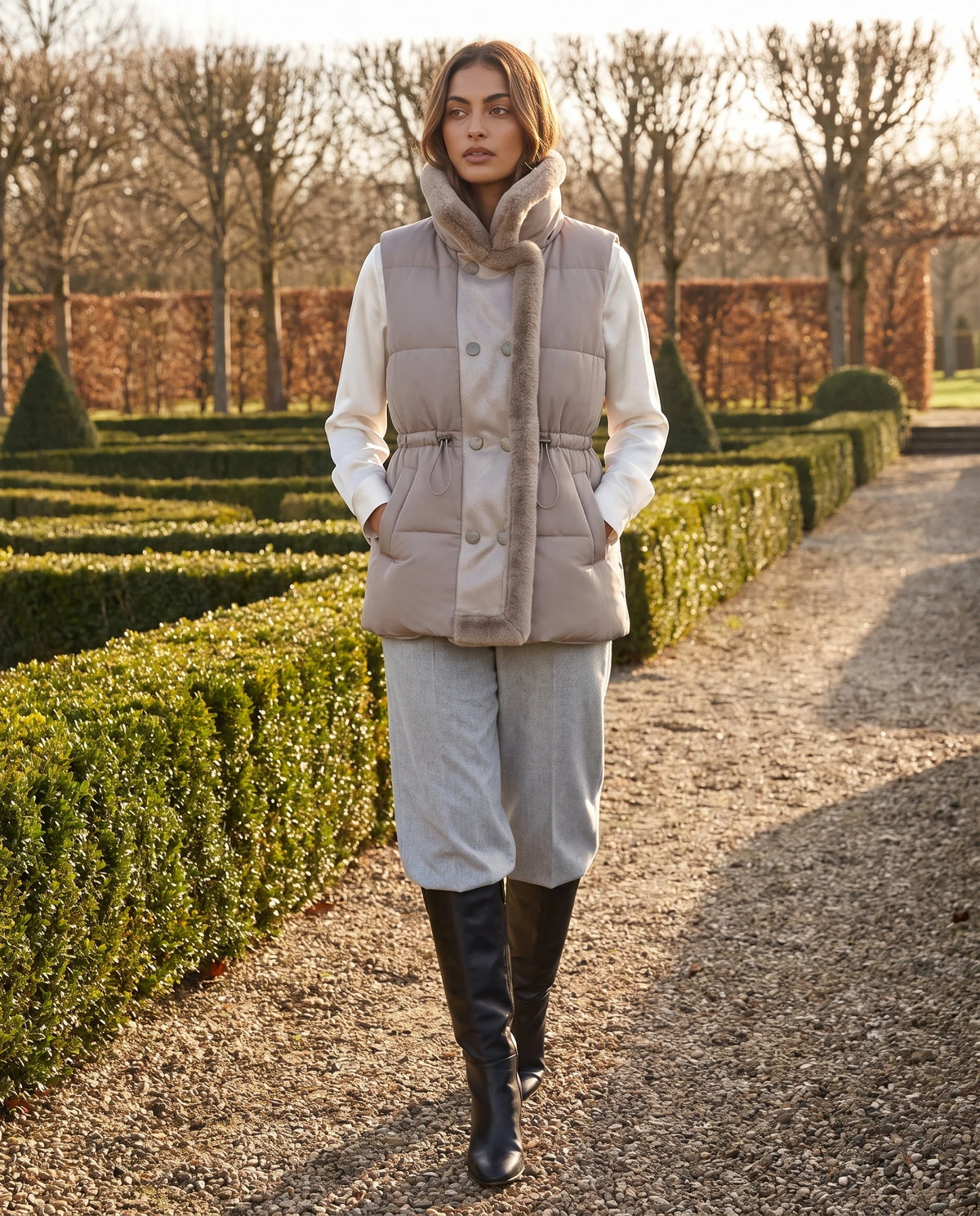 Gilet sans manches à col fausse fourrure Zélia Taupe Clair, porté col relevé dans une allée de jardin à la française