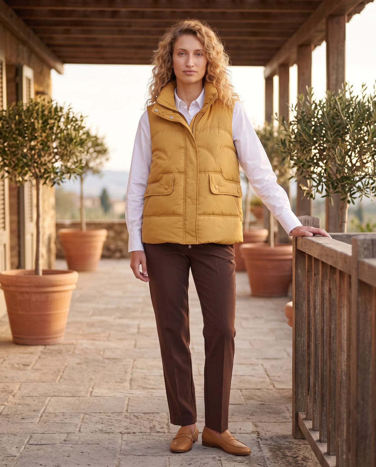 Gilet matelassé court à col montant Sylvie en Moutarde, porté sous une pergola en bois, entre des oliviers en pot