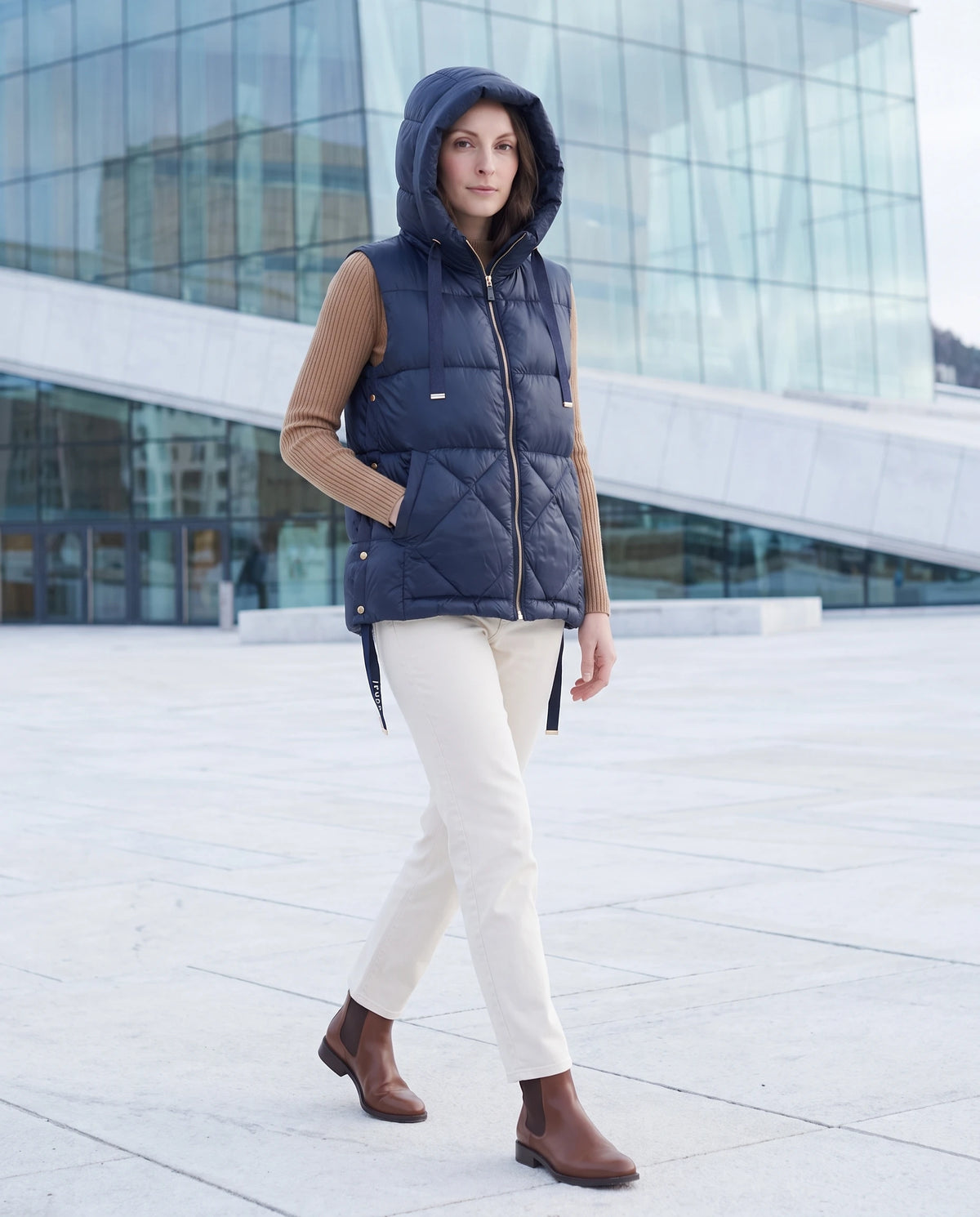 Gilet matelassé à capuche et grandes poches Corinne en Bleu Marine, capuche relevée, sur une esplanade devant un bâtiment vitré