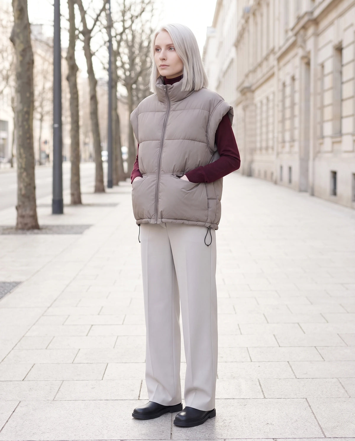 Gilet matelassé sans manches à poches plaquées Anceline en Gris Taupe, porté dans une rue de pierre claire, mains dans les poches