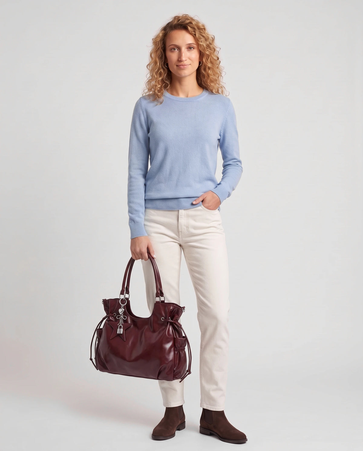 Grand sac à main hobo de cours bandoulière à breloque étoile Lyra coloris Bordeaux, porté à la main par ses deux anses, sur un pull fin bleu poudré et un jean droit écru