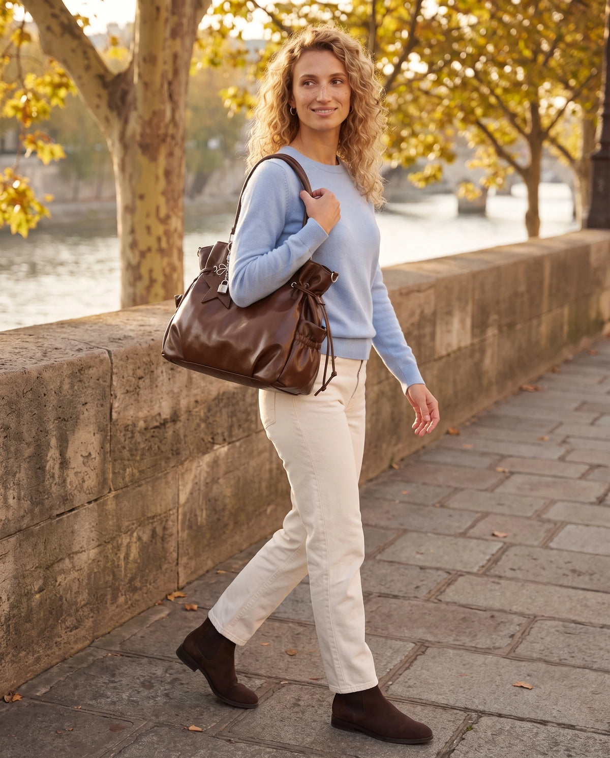 Grand sac à main hobo de cours bandoulière à breloque étoile Lyra coloris Marron Châtaigne, porté à l'épaule sur un quai de pierre au bord de l'eau à l'automne