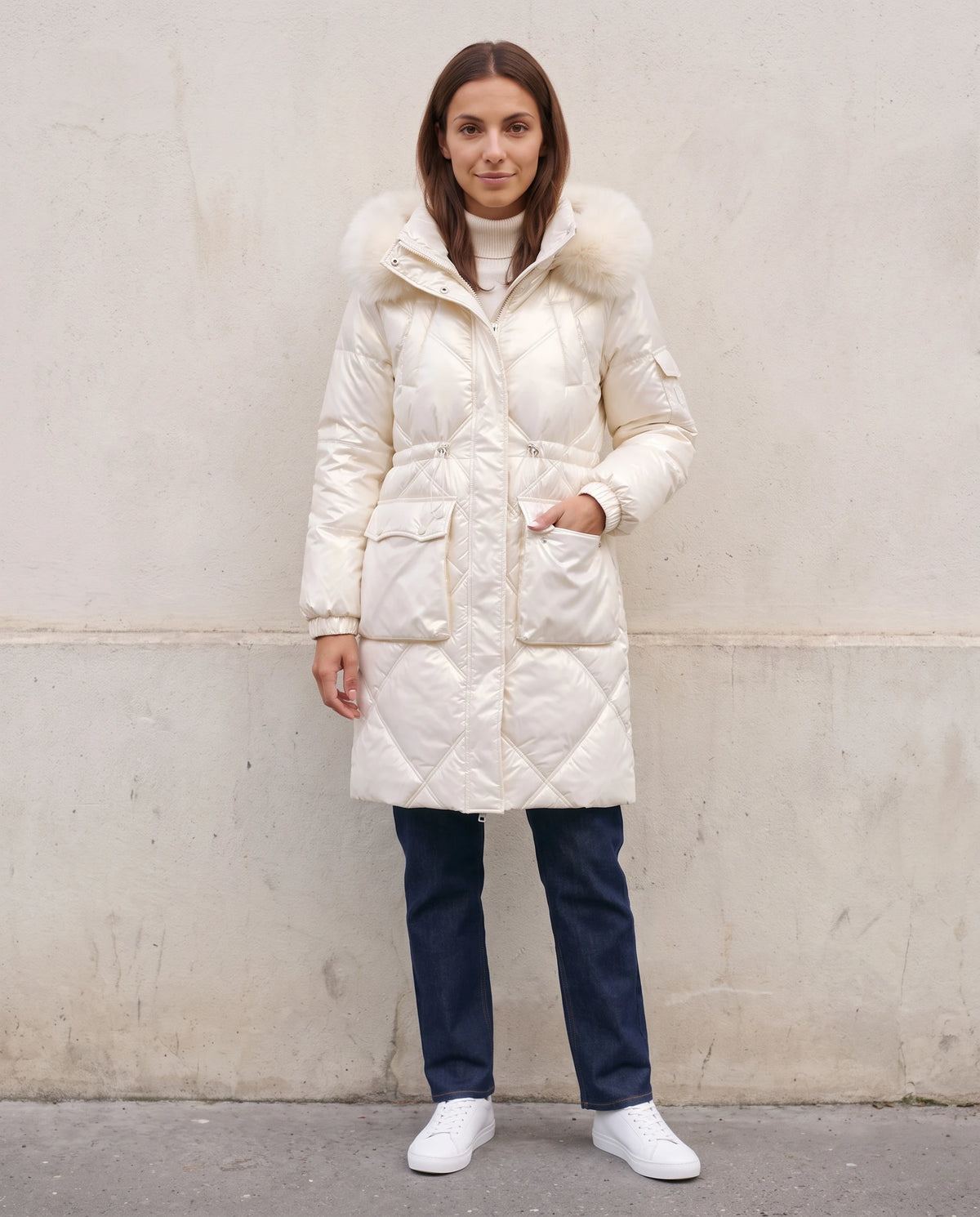 Parka mi-longue brillante à capuche fausse fourrure Vanessa en Blanc Cassé, portée devant un mur de pierre claire