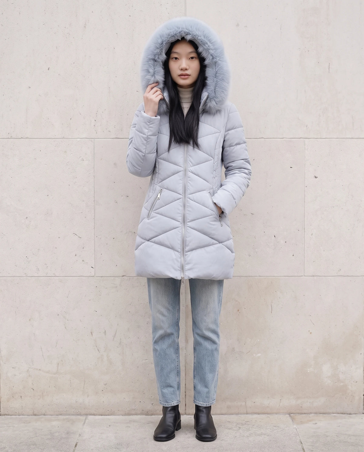 Parka mi-longue cintrée à capuche fausse fourrure Paola en Bleu Gris, portée devant un mur de pierre claire, capuche relevée