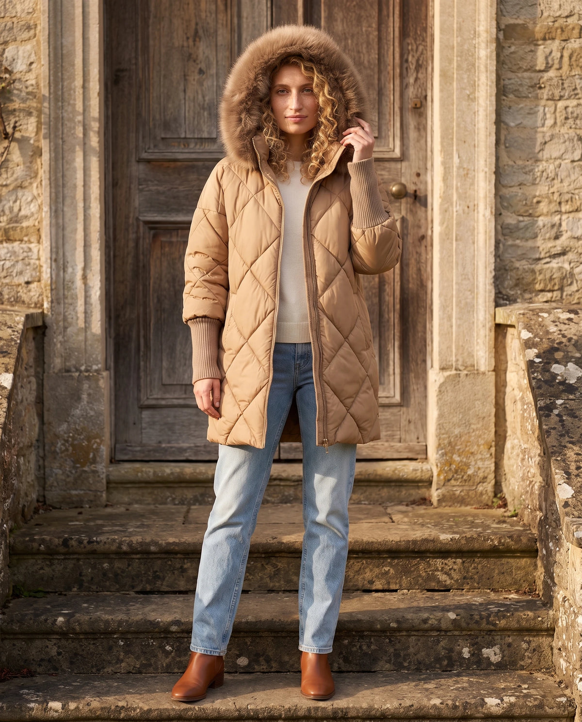 Parka mi-longue matelassée à manches tricot Ava en Camel, portée sur le perron de pierre d'une maison de campagne, capuche relevée
