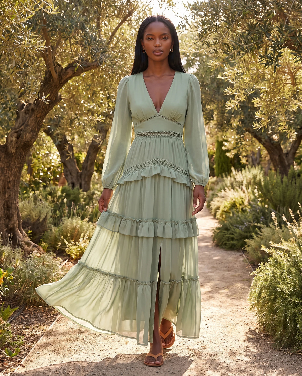 Langes Boho-Kleid mit Volants und Häkelborten Inaya in Mandelgrün, unter den Olivenbäumen eines Gartens getragen