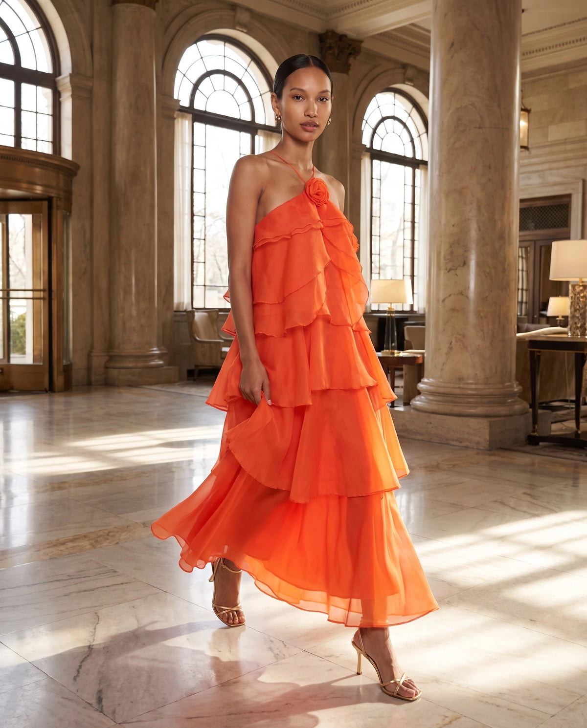 Robe longue halter en organza à volants étagés Iliana en Orange, portée dans un hall d'hôtel en marbre