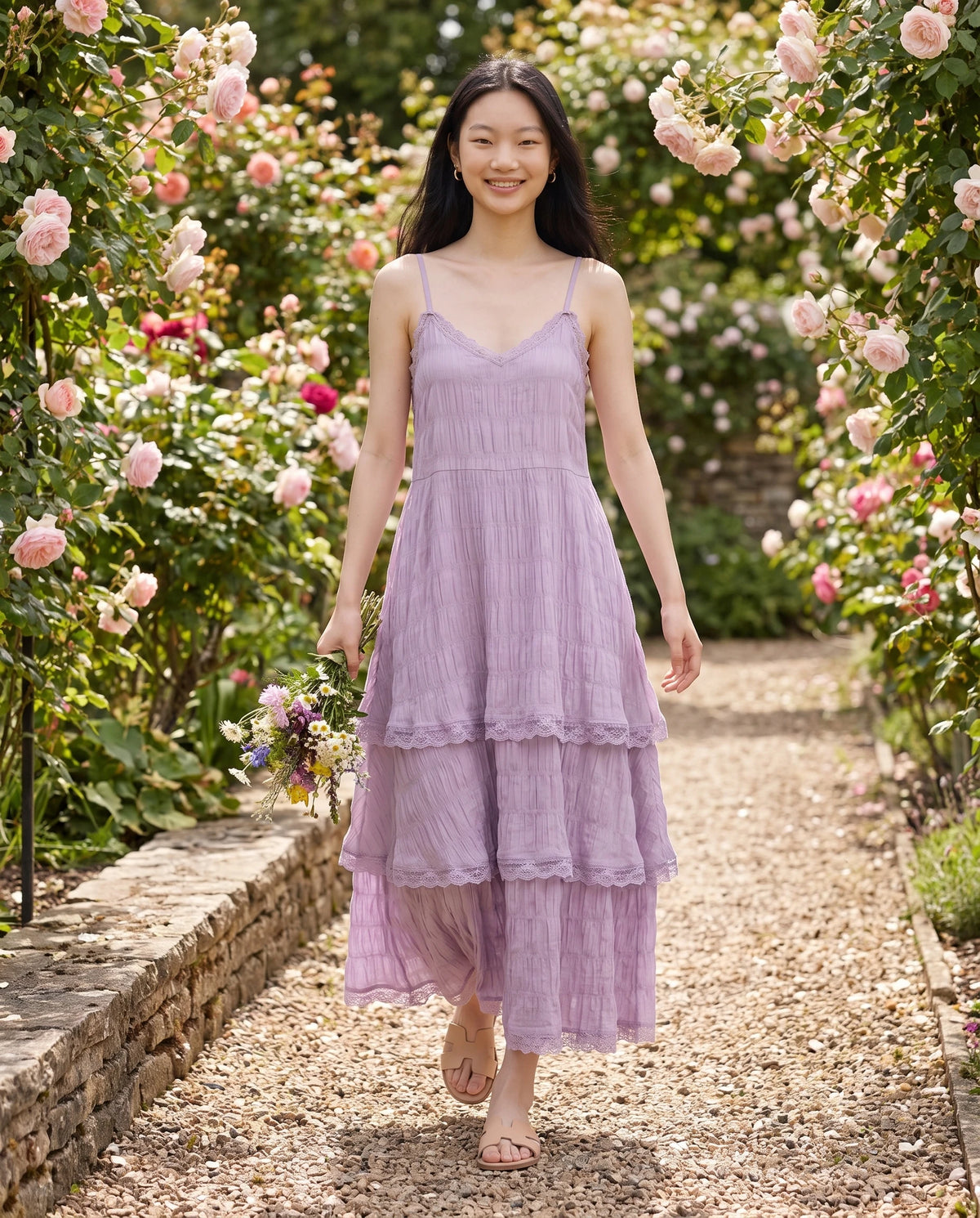 Robe longue à volants étagés et dentelle Elina en Lilas, portée sur une allée de roseraie