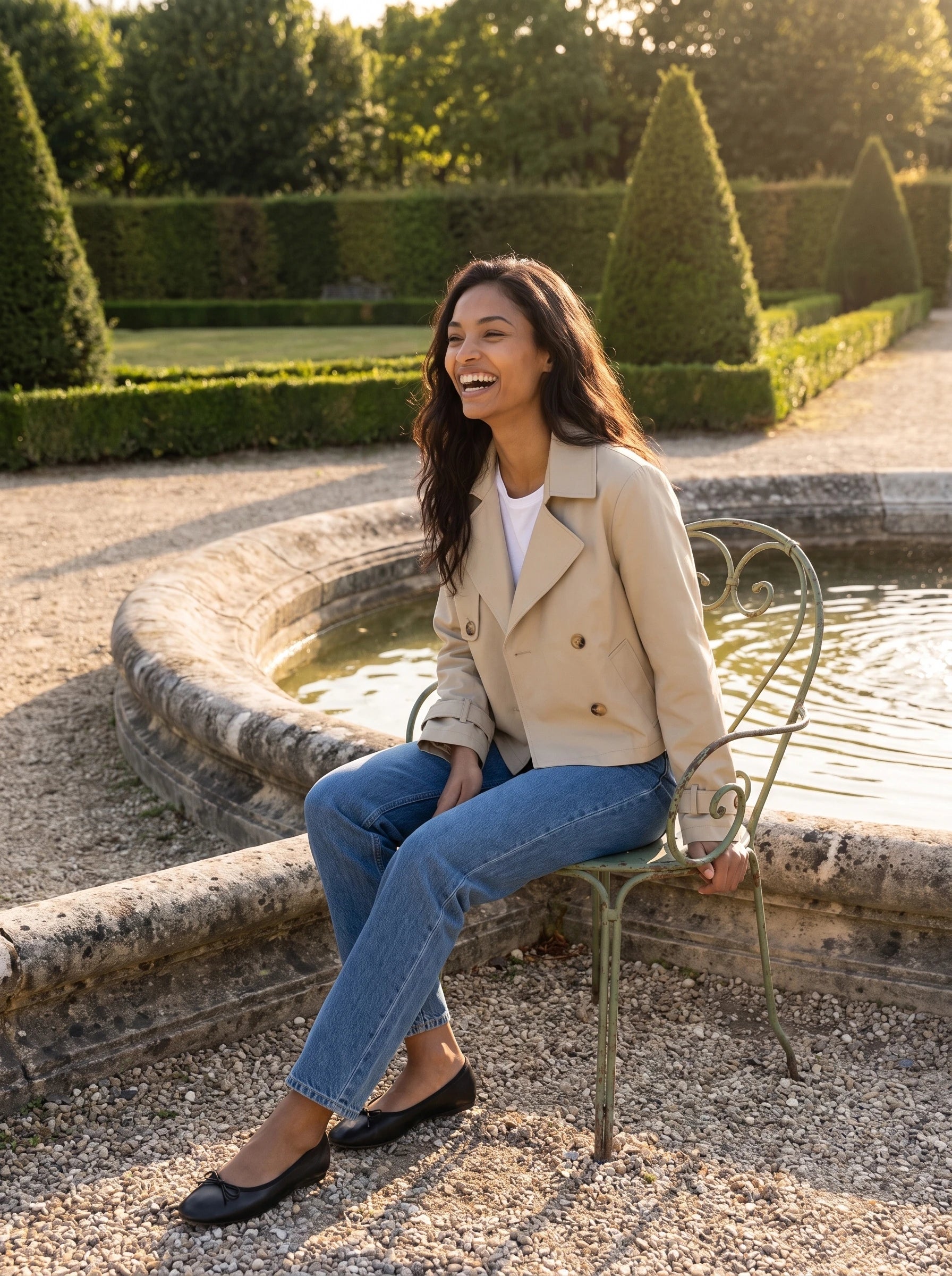 Trench court croisé beige Fernande, porté près d'une fontaine de jardin à la française