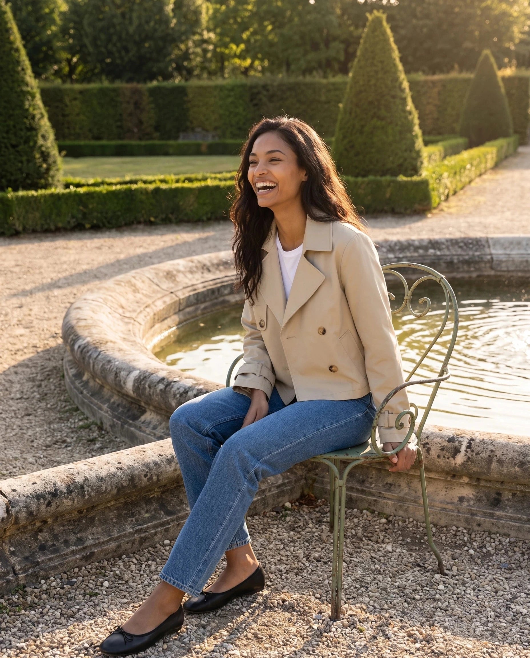 Trench court croisé beige Fernande, porté près d'une fontaine de jardin à la française