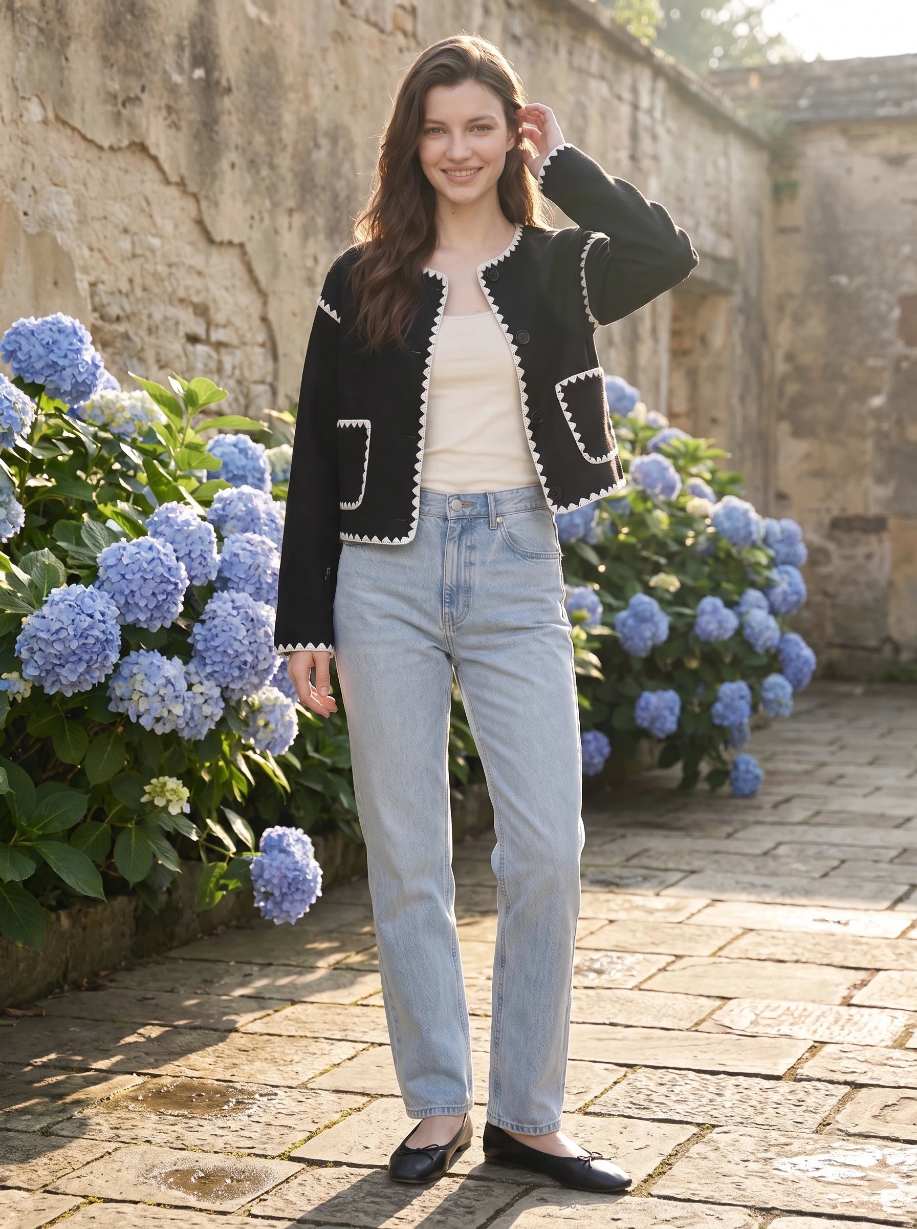 Veste courte festonnée noire Huguette, portée dans une cour aux hortensias bleus