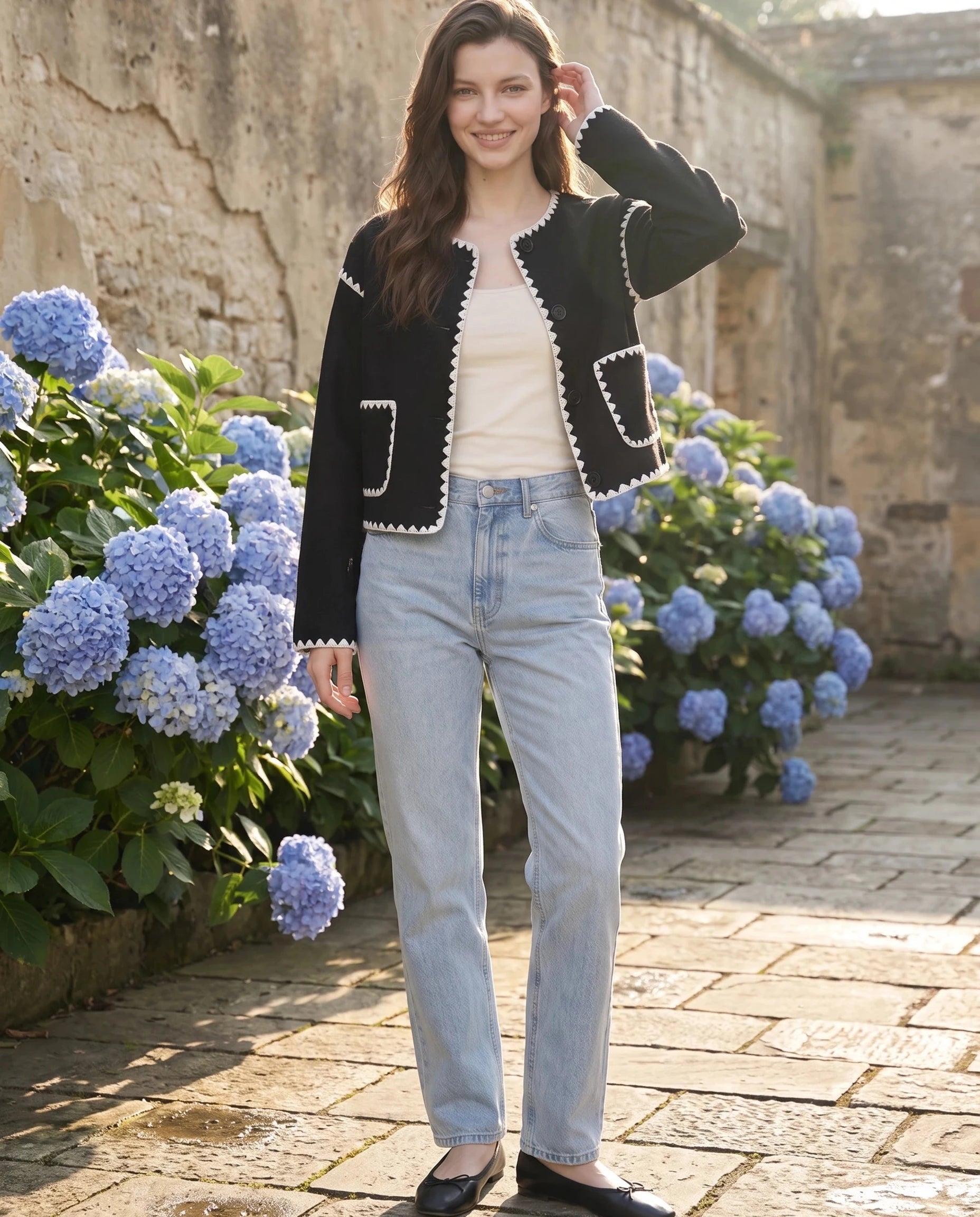 Veste courte festonnée noire Huguette, portée dans une cour aux hortensias bleus