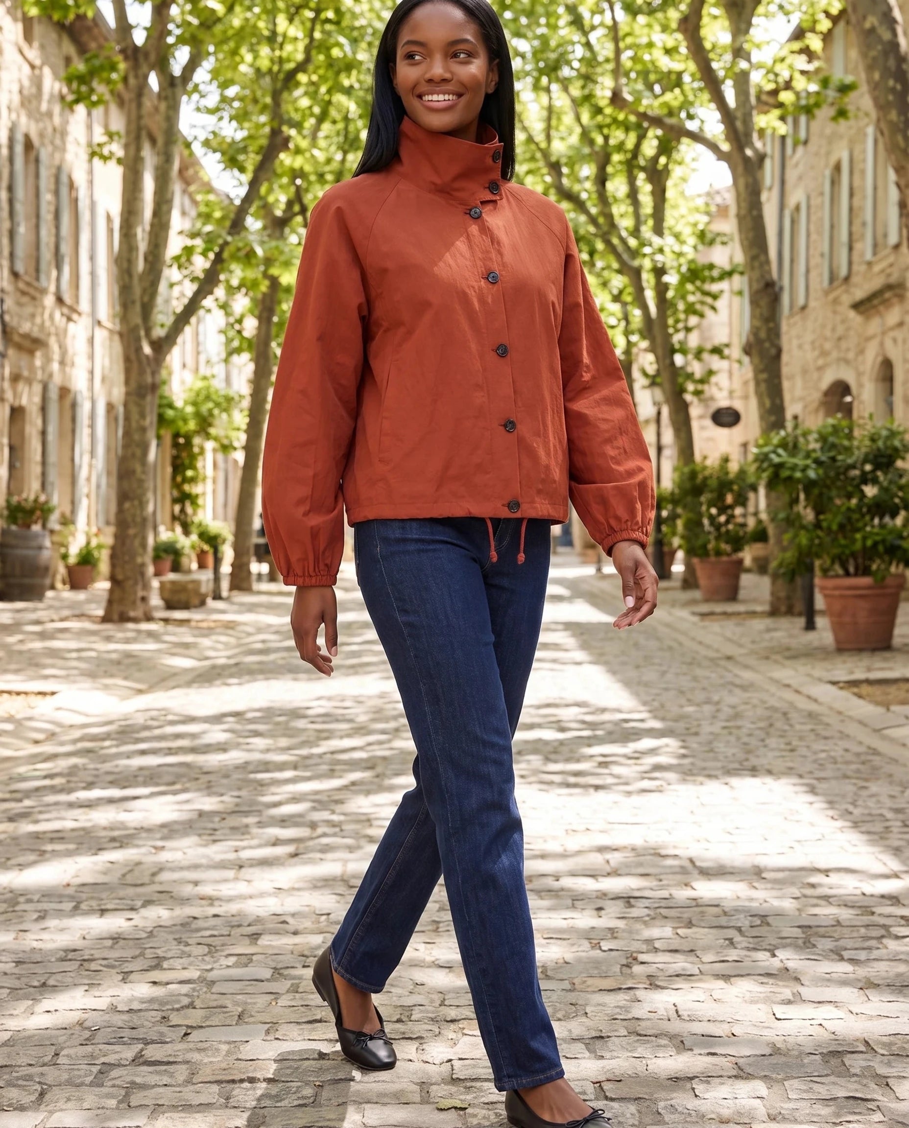 Veste légère rouge Euphrasie, portée dans une allée de tilleuls d'un village
