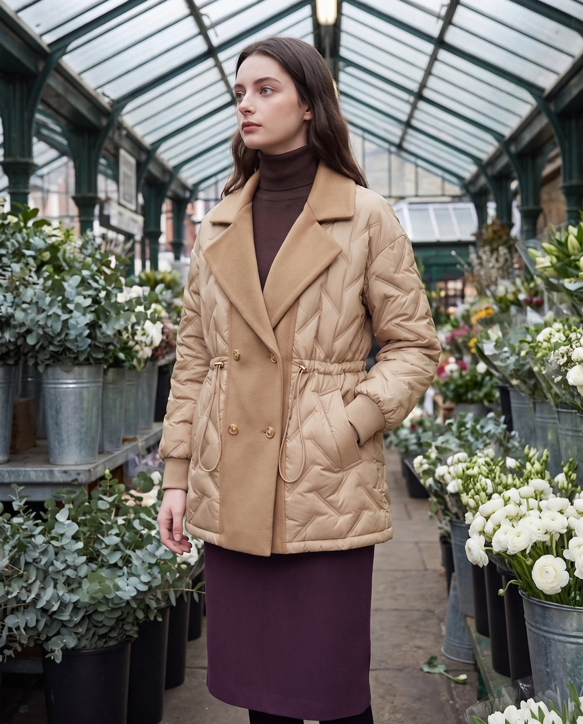 Veste matelassée mi-longue à grand col tailleur Géraldine en Beige Sable, portée sous la verrière d'un marché aux fleurs