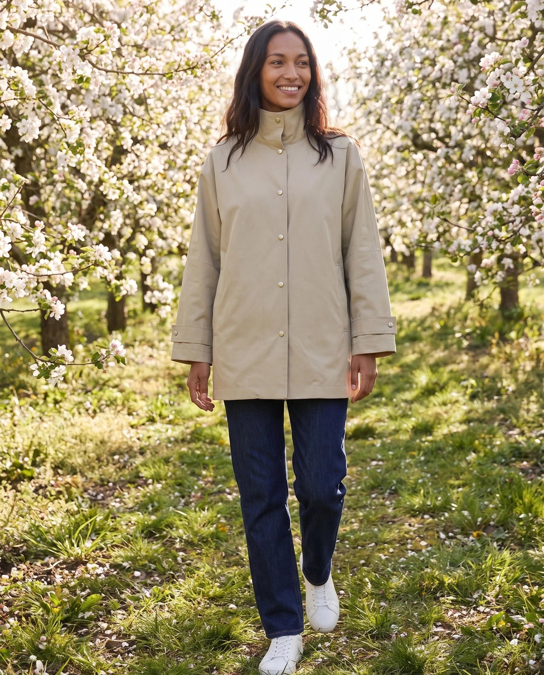 Veste mi-longue beige sable Mauricette, portée dans un verger de pommiers en fleurs
