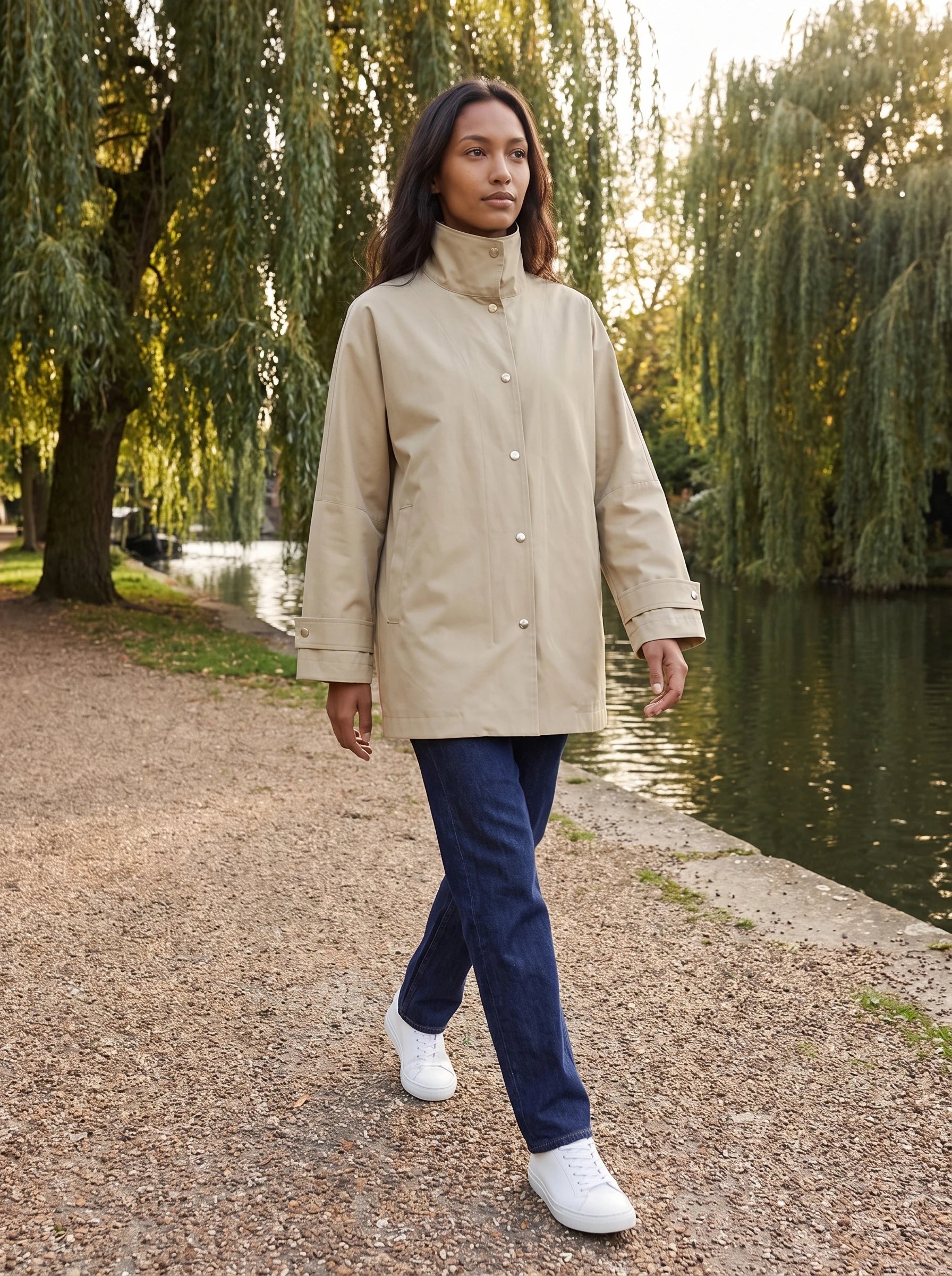 Veste mi-longue beige sable Mauricette, portée le long d'un canal bordé de saules