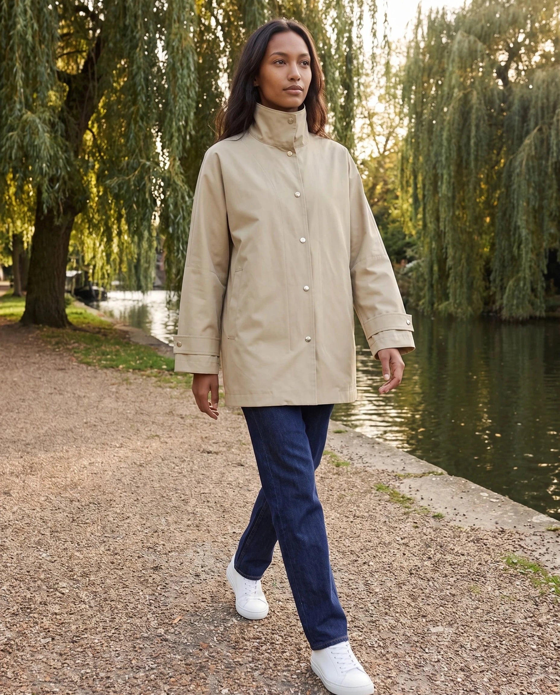 Veste mi-longue beige sable Mauricette, portée le long d'un canal bordé de saules
